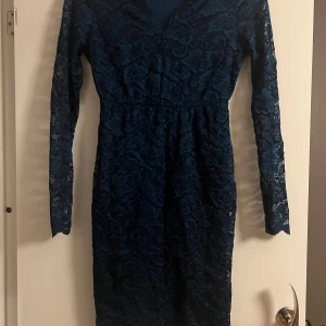 Mörkblå spetsklänning - Elegant mörkblå klänning i spets med långärmad design. Klänningen har en v-ringad hals och en figurnära passform som framhäver silhuetten. Perfekt för en stilfull kväll.