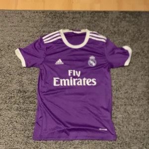 Lila Real Madrid fotbollströja - Säljer en lila Real Madrid fotbollströja från Adidas med 'Fly Emirates' tryck på framsidan och 'Ronaldo 7' på baksidan. Tröjan har korta ärmar och vita detaljer vid krage och ärmslut. Perfekt för fans av Ronaldo och Real Madrid! ⚽️