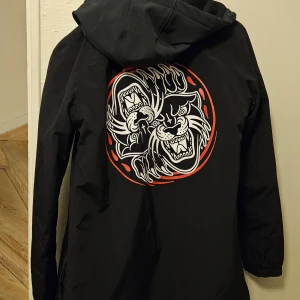 Svart jacka från Lurking Class storlek S - Snygg svart snowboard jacka från Lurking Class med ett coolt tryck av en tiger på ryggen. Jackan har en huva och stängs med dragkedja och knappar. Perfekt för dig som vill ha en unik stil.