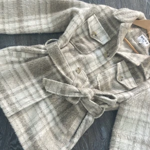 Rutig kappa från NA-KD - Säljer en snygg rutig kappa från NA-KD i beige och grå toner. Den har ett bälte i midjan och knappar framtill. Perfekt för höst och vinter med sin stilrena design och långa ärmar. Ett måste för garderoben! 