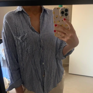 Blåvit randig skjorta från Zara - Snygg blåvit randig skjorta från Zara med klassisk krage och knappar framtill. Skjortan har en bröstficka och långa ärmar, perfekt för en avslappnad stil. Tillverkad i linnematerial.