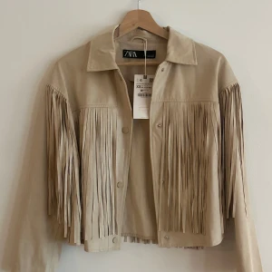 Beige mockajacka med fransar från Zara - Snygg beige mockajacka från Zara med fransar längs ärmar och rygg. Jackan har en klassisk krage och knappar framtill. Perfekt för en trendig look. Storlek xs