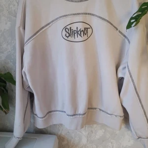 Vit Slipknot sweatshirt - Säljer en vit sweatshirt med Slipknot-logga framtill. Tröjan har långa ärmar och kontrasterande sömmar som ger en cool look. Perfekt för fans av bandet!