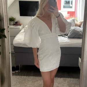 Student klänning - Aldrig använd och prislappen är kvar. Säljer en vit klänning med puffiga trekvartsärmar och v-ringning. Klänningen har en tight passform och är tillverkad i ett broderat material. Passar perfekt som studentklänning