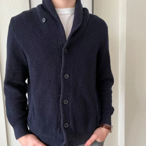 Mörkblå stickad kofta med knappar - Säljer en stilren mörkblå stickad kofta med knappar framtill. Koftan har en snygg krage och långa ärmar, perfekt för kyligare dagar. Den har en texturerad yta som ger en extra touch av elegans. Passar bra till både vardag och mer uppklädda tillfällen.