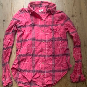 Rutig rosa skjorta från Hollister - Säljer en snygg rosa rutig skjorta från Hollister. Skjortan har långa ärmar och knappar framtill. Perfekt för en avslappnad stil med en bröstficka för extra detalj.
