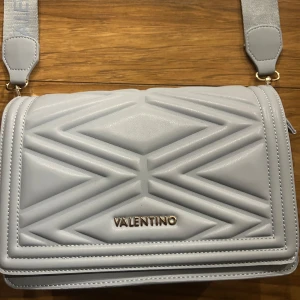 Ljusblå axelväska från Valentino - Snygg ljusblå axelväska från Valentino med quiltad design och justerbar axelrem. Väskan har en dragkedja för säker förvaring och ett elegant märke framtill. Perfekt för att ge din outfit en lyxig touch. Köpt på Sellpy för 650. Använd 1 gång, säljer för 600.