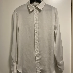 Vit långärmad skjorta - Säljer en klassisk vit långärmad skjorta. Skjortan har en stilren design med knappar framtill och vid ärmsluten. Perfekt för både formella och avslappnade tillfällen. Den är redo att bäras till nästa event! Aningen urtvättad annars fin