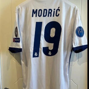 Vit Real Madrid fotbollströja med Modrić 19 - Säljer en vit Real Madrid fotbollströja från Adidas med Modrić och nummer 19 på ryggen. Tröjan har korta ärmar och är dekorerad med flera märken, inklusive FIFA och UEFA Champions League. Den har en klassisk krage med knappar och Fly Emirates som sponsorlogga på framsidan.