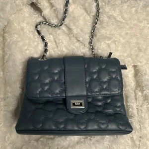 Blå quiltad axelväska med hjärtmönster - Säljer en superfin gråblå axelväska med quiltat hjärtmönster som är köpt i Kroatien förra året! Den är använd fåtal gånger och är i bra skick förutom slitningen som ni ser på sista bilden, men det syns knappt och går att laga enkelt!💙