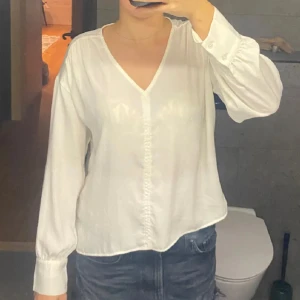 Vit blus från Vero Moda - Säljer en stilren och elegant vit blus från Vero Moda. Den har en v-ringad design och långa ärmar, perfekt för både vardag och fest. Materialet är lätt och bekvämt, vilket gör den idealisk för vår och sommar. Passar bra till både jeans och kjol!
