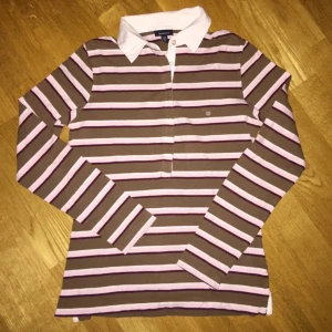 Randig pikétröja från Gant. M - Säljer en randig pikétröja från Gant i brunt och rosa. Stl M. Bommulstrikå. Den har en klassisk vit krage och långa ärmar! Slits i sidorna. 