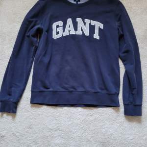 Säljer en snygg mörkblå sweatshirt från GANT i storlek 2XL. Tröjan har ett stort, vitt GANT-tryck på bröstet och är perfekt för en avslappnad stil. Den är långärmad och gjord i ett mjukt material som är superbekvämt. Perfekt för höst och vinter!