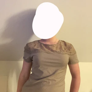 Beige topp med spetsdetaljer - Säljer en snygg beige topp med korta ärmar och fina spetsdetaljer över axlarna och övre delen av ryggen. Perfekt för både vardag och festliga tillfällen. Toppen har en normal passform och är superbekväm att bära.