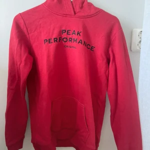 Röd hoodie från Peak Performance - Säljer en snygg röd hoodie från Peak Performance. Den har en dragkedja vid halsen och ett tryck med märkesnamnet på bröstet. Perfekt för kyliga dagar och passar till det mesta i garderoben. Superbekväm och stilren!