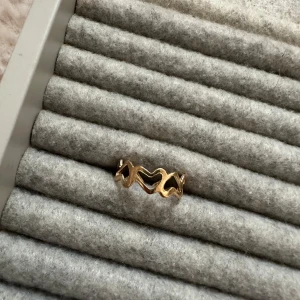 Guldfärgad ring med vågig design - Säljer en snygg guldfärgad ring med en unik vågig design. Perfekt för att ge en stilren touch till din outfit. Ringen är i nyskick och passar till både vardag och fest. En riktig statement piece för smyckesälskare!