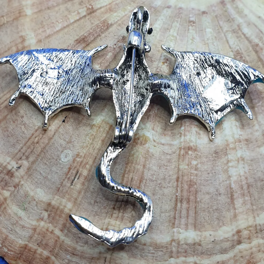 En brosch föreställande en drake, storlek ca 7x6cm.  Silverfärgad metall med vacker ytstruktur och emaljering samt kristaller.  Grymt snygg brosch, oanvänd, nyskick.. Accessoarer.