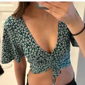 Grön blommig topp - Divided - Säljer en superfin grön topp med vita blommor från Divided. Toppen är croppad och har en snygg knytning framtill. Perfekt för sommardagar eller en festlig kväll! Mycket fint skick, inga defekter. Storlek xs. 🌸