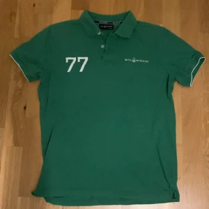 Grön pikétröja från Sail Racing - Säljer en snygg grön pikétröja från Sail Racing i storlek medium. Tröjan är i bomull och har korta ärmar med vita detaljer. Den har en klassisk krage och knappar framtill. Perfekt för en avslappnad stil eller en dag på stan!