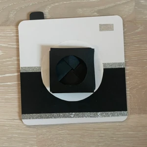 Kort - Det här är en kreativ handgjord kortdesign som ser ut som en kamera. Kortet är fyrkantigt med en vit och svart färgkombination och har glittriga detaljer. Det finns en roterande del i mitten som avslöjar ett meddelande 'For you' med en söt illustration av hus och träd. Perfekt för att ge en personlig touch till en present.