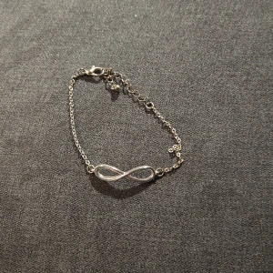 Silverfärgat armband med oändlighetssymbol - Snyggt och stilrent armband i silverfärg med en oändlighetssymbol i mitten. Perfekt för både vardag och fest. Armbandet har en justerbar kedja med en liten pärldetalj vid låset. Enkelt att matcha med andra smycken för en komplett look.