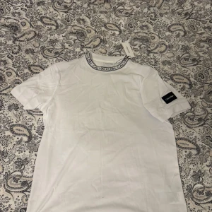 Vit t-shirt från Calvin Klein - Säljer en vit t-shirt från Calvin Klein i nyskick. Den har en cool logga runt halsen och en patch på ärmen. Perfekt för en avslappnad stil och passar till alla tillfällen. T-shirten är i bomull och har en normal passform.