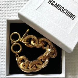 Moschino x HM armband - Kollar intresset på detta galet snygga guldplateradd armbandet fråndet exklusiva samarbetet mellan moschino och hm (limited edition) ❤️ Aldrig använt så precis i nyskick! Det är platerat med guld 🥰 Sjukt bra kvalitet och känns dyrt o lite tungt