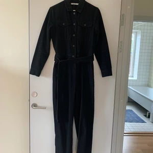 Jumpsuit i svart jeans - Jumpsuit / boilersuit i svart jeanstyg. Normal passform på överdelen (sitter inte jättetight), nederdelen sitter som mom jeans (smalnar av lite nedan för knäna men sitter inte tight). Skärp i midjan. Fickor fram på byxorna. Superfint skick, inte mycket använd. Material 100% bomull 