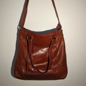 Skinnväska - Rymlig och trevlig väska i skinn. Märket är Vezzano och den har en brunröd färg. Går att bära med både den långa remmen och som en handväska med de korta. Rymmer mycket och har små fack på insidan. I gott skick!