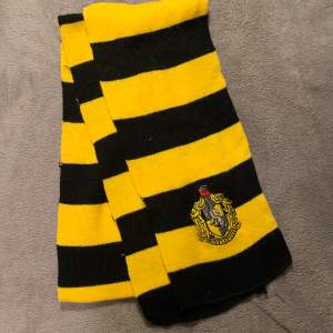 En randig halsduk i svart och gul med en embroiderad Hufflepuff logga. Oanvänd. Från Science Fiction Bokhandeln 80kr + frakt (cirka 50kr)