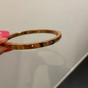Stelt armband med detaljer i form av nyckelhål och nyckel. Öppnas genom knäpp-mekanism (bild 2, nyckel) . Roséfärg