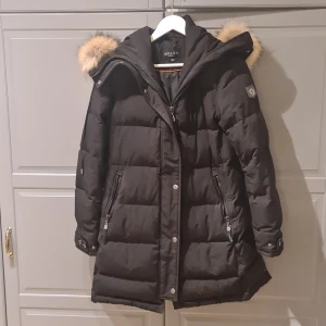 Hollies  subway jacket - Svart, figursydd dunjacka i längre modell från hollies. Luva med avtagbar pälskrage i beige naturpäls, 2 fickor fram med dragkedja. Använt skick