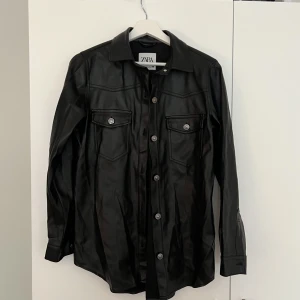 SKINN SKJORTA ZARA - Supersnygg svart skinn skjorta i fejkskinn från Zara! Lite skrynklig på bild men har legat nedlagd och oanvänd!  Använd fåtal gånger och är väldigt fint skick! Super snyggt nu till våren! 