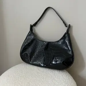 En svart handväska från Gina tricot med glansigt material. Materialet är ”Krokodilskinn” 