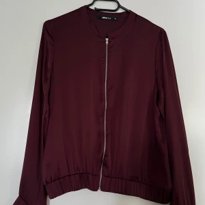 Tunn jacka Gina Tricot vinröd 36 - Vinröd tunn jacka (bomber stil) från Gina Tricot, storlek 36. Tunnt material, klippt av lappen men misstänker polyester. Stängs med dragkedja fram. Jackan är mycket god skick.   Mått: Längd ca 60 cm Byst ca 50 cm Midja ca 50 cm Ärmlängd mätt från axe
