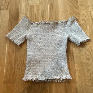 En beige offshoulder topp med korta ärmar och volangdetaljer vid ärmslut och nederkant. Toppen har en rynkad textur och är tight passande.