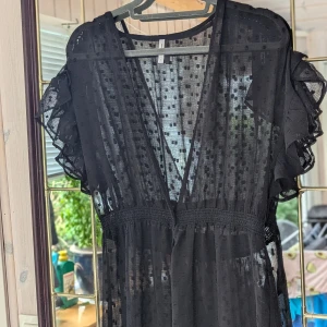 Svart klänning från Vero Moda/lång kimono  - En elegant svart kimono från Vero Moda med ett prickigt mönster. Klänningen har korta ärmar med volangdetaljer och en v-ringad hals. Den är tillverkad av 100% polyester och har en smickrande passform med en markerad midja.mycket fint skick.