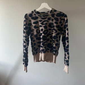 Leopardmönstrad stickad tröja från Dilvin - En bekväm och stilren ulltröja från Dilvin med ett leopardmönster i svart och beige. Tröjan har långa ärmar och ribbstickade muddar vid ärmslut och nederkant.