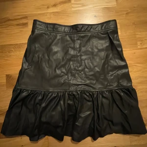 Kjol - Svart minikjol i läderimitation med volangkant. Stängs med dragkedja som sitter vid höften. Använd några gånger men i fint skick.