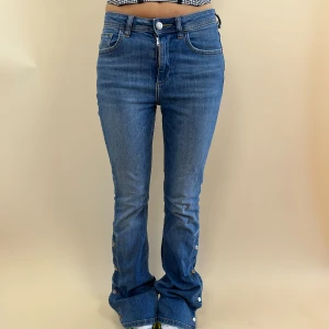 Blå jeans med knappar - Midjemåttet är 75cm, dock är midjan strechig!! Innerbenslängden är 85cm. Utsvänga jeans med knappar längst benen som också går att knäppa upp. Modellen är 165cm