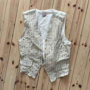 Beige rutig väst - En beige rutig väst med knappar framtill och två fickor. Västen har en vit baksida med knytning.