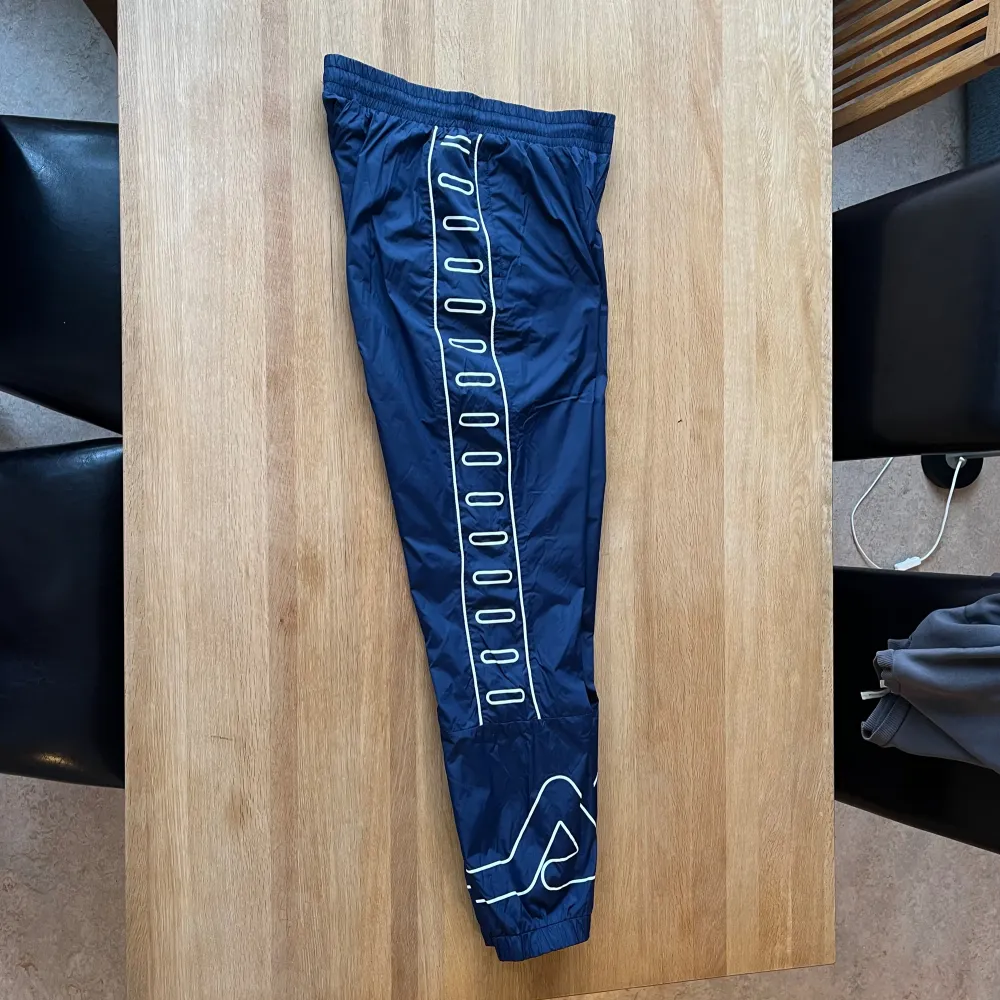 Mörkblåa byxor med vita detaljer från Fila. Vindtätt tyg, två sidofickor med dragkedja, resår och snörning i midjan. Använda ett fåtal gånger.. Jeans & Byxor.