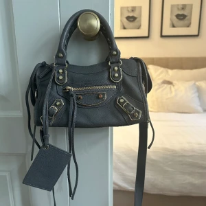 Balenciaga mini city bag  - Säljer min balenciaga city bag💫 Fint skick förutom att niten där bak är borta, har en säkerhetsnål där nu men jätte enkelt att fixa om man vill det. Spegeln har jag tagit bort, även de går o sätta i en ny.  Grå med guldiga detaljer🤍⚡️
