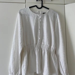 Vit blus - En vit blus med en dragkedja bakom ryggen och små prickiga detaljer. Den är mellan lång blus som sitter vid midjan och som är löst nedåt.