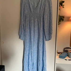 Klänning - Blå långklänning med vit blommor från Nelly. Använd fåtal gånger och i nyskick. Storlek 38