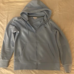 Lacoste hoodie zip - Ljusblå hoodie perfekt till att matcha med ljusa outfits till sommaren. Köpt i mars och använd fåtal gånger. 