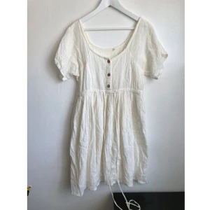 Vit kortärmad miniklänning, 34/36 XS/S, vintage, retro, bohemisk, boho, student - Vit miniklänning i krinklad bomull, knappar fram, dragsko i midjan. Ytslitage finns samt fläckar. Mått i cm: bröstvidd 45; längd fram från axeln 88; ärmlängd mätt från halsen 23.