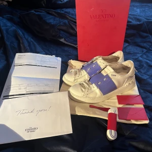 Valentino garavani skor - Ett par vita och lila Valentino Garavani sneakers i skinn. Skorna har snörning och en bred lila/blå rem över mitten. De kommer med originalförpackning och tillbehör. det finns även KVITTO, köpta på miinto