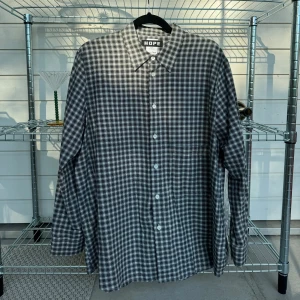 Hope, Elma shirt - grå/vit/svart check - Strl: W34/M42 Inga ”synliga” defekter, däremot så har tyget bakom sista knappen gått upp. Inget som påverkar funktionen eller knappen, då knappen sitter fast på ett annat lager. Vet ej om bilden visar tydligt hur det har gått upp. Är bakom knappen
