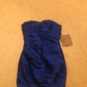 Kungblå axelbandslös klänning - En elegant kungblå axelbandslös klänning med en åtsittande passform. Klänningen har en vacker detalj med pärlor och paljetter längs den övre kanten och en knutdetalj framtill. Materialet är glansigt och ger en lyxig känsla.